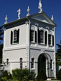 Un petit blanc.  maison à peu près carrée.  Il est richement décoré de statues au sommet du pignon du toit, de parements travaillés en forme de pilier et d'un fronton de pignon avec moulures dentelées.  Toutes les garnitures sont blanches à l'exception des volets, qui sont noirs.