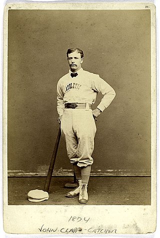 A baseball player is shown standing in his baseball uniform, leaning on the end of a baseball bat.