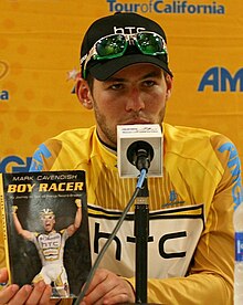 Cavendish sitting in front of a microphone holding a copy of his book Boy Racer