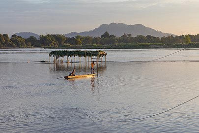 Mekong pirogue at sunset with two men working at a power line to connect a partially submerged shelter in the 4000 islands (Si Phan Don) near Don Puay, Laos