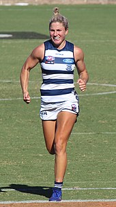 A female athlete with light hair wearing a sleeveless guernsey and shorts jogs on the grass surface of the playing arena.