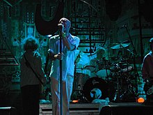 Cantor masculino em mangas de camisa e calças brancas, com uma banda atrás dele em um pequeno palco.