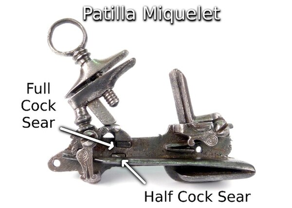 The patilla style miquelet lock fully cocked. An extension of the cock is resting on and restrained by the upper of the two horizontal sears protrudin