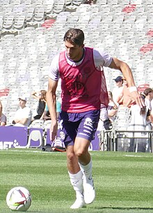 Stijn Spierings en tenue d'échauffement (short violet, chasuble rose et chaussettes blanches) s’apprête à tirer dans un ballon.