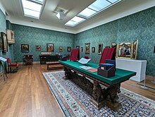 A large room with skylights and wooden floors, displaying a range of paintings on the walls and on tables