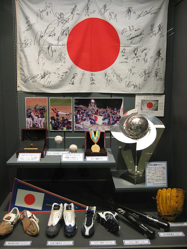 Photo en couleur d'une vitrine, au premier plan des chaussures, gants et battes de baseball, au deuxième rang des médailles et un trophée, à l'arrière plan un drapeau blanc et rouge