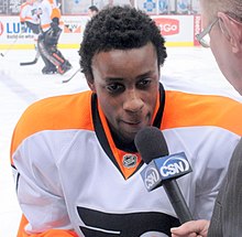 A Black man in an orange-and-white hockey jersey speaks into a microphone.