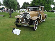 Ein braunes Auto im Stil der 1920er Jahre, das nach vorne zeigt, ist auf Gras links abgebildet