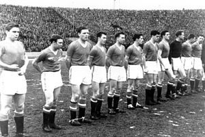 A black-and-white photograph of a football team lining up before a match. Eleven men in old-fashioned association football attire stand in a line: ten wear dark shirts, white shorts and black socks, and the other wears a still darker shirt. Behind the players can be seen one of the enormous open stands of an East European-style soccer stadium, filled to the brim with spectators.
