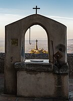 Rank: 23 View of the stone cross Creu d'Es Picot on the beside mountain Puig des Milá, framed by the drinking fountain at the Sanctuary of Sant Salvador