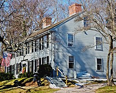A blue colonial two story house with black shutters. Over the front door is a flag pole holding an American flag.
