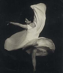 An artistic photograph of a woman dancing, in mid-spin; she is wearing a light-colored lightweight dress and has bare feet.