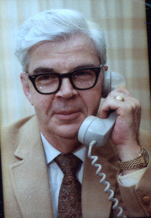 Un homme aux cheveux blancs, portant des lunettes à monture de corne et un costume beige, tient un téléphone dans sa main gauche.