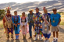 Members of the Jicarilla Apache tribe gather at the Great Sand Dunes to share traditional clothings, crafts, stories, and dances in July of 2019. Jicarilla Apache Mundo Dancers, Dunes in Background (48390690046).jpg