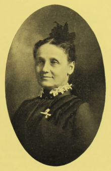 B&W oval portrait photo of a middle-aged woman with her hair in an up-do, wearing a dark blouse with a white frilly collar, and a white ribbon pinned on the blouse.