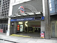 An entrance in a larger building under a sign reading "MONUMENT STATION" reveals banisters leading down. A woman is walking out of the entrance