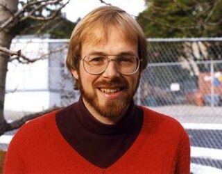 Portrait d'un homme blond, barbu, portant des lunettes, qui sourit en regardant l'objectif.