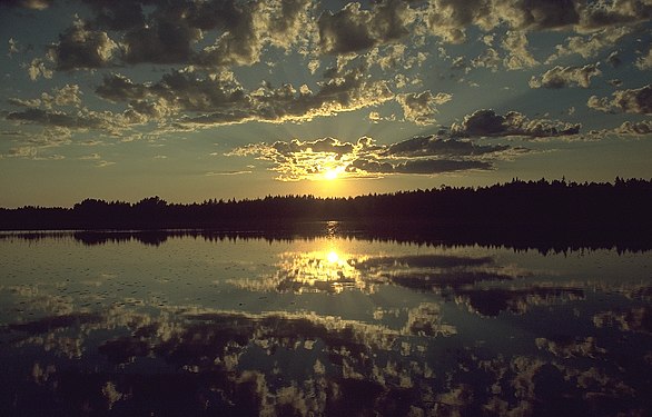 Sun setting over forest and lake; sun and clouds reflecting in lake.