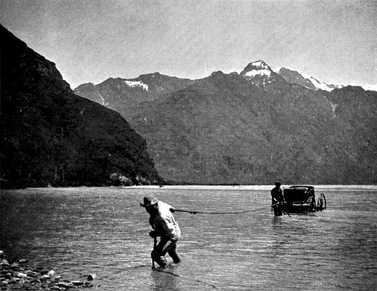 A four-wheeled carriage in a calm river is pulled ashore with a rope by two men. There is a bush-covered hill to the left and mountains with a scattering of snow near the peaks in the background.