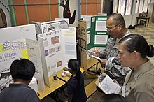 Science fair project display US Navy 100416-N-5539C-006 Construction Electrician 3rd Class Jill Johnston and Lt. Col. Johnny Lizama listen to third-graders from Harry S. Truman Elementary School explain their science projects.jpg