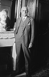 Un homme en costume se tient debout, main dans la poche de pantalon, devant un buste et un miroir.