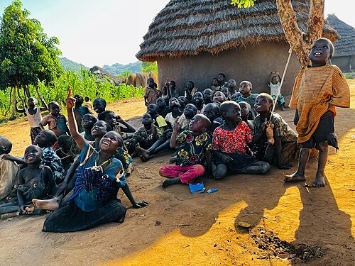 Children gathered outside a traditional thatched roof house in Karamoja region while gazing at the flying drone in the clear sky by Sandra Aceng