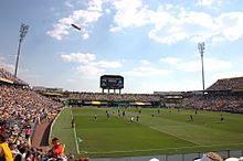 Un estadio de fútbol con un campo de césped, multitudes en las gradas, un marcador distante y un dirigible volando por encima de su cabeza durante el juego.
