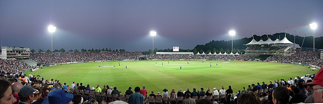 Panorama of a heavily populated cricket ground at night.  A large stand is on the right-hand side of the pitch and the scene is illuminated by four floodlights.