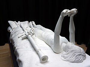 Foto of life size statuario marble sculpture with a black background. The sculpture is of a bed, with a naked young woman laying down, taking a selfie with a photo camera. Beside her on the bed lay large military weapons