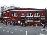 A red-bricked building with a rectangular, blue sign reading "HAMPSTEAD STATION" in white letters and people in front all under a white sky