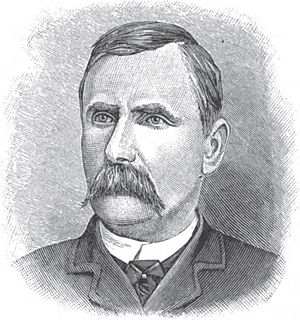 A man with dark hair and a dark mustache wearing a white shirt, black tie, and black jacket