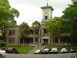 Photograph of the Edificio José de Diego, an imposing, two-story building on top of a short rise, flanked by large trees and with a tall clock tower centered on the symmetrical facade