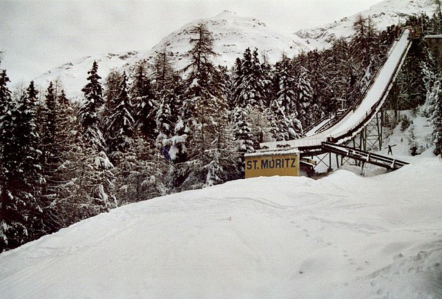 Image d'un tremplin de saut à ski avec en bas l'inscription "St. Moritz" et derrière lui, des arbres enneigées.
