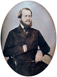 Photographic half-length portrait of a seated bearded man dressed in a dark, double-breasted coat with his right hand tucked inside the front