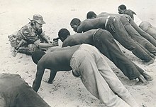 Photographie d'hommes noirs torses nus en équilibre sur les mains et les pieds. Un soldat blanc pointe son pistolet sur le front de l'un d'eux