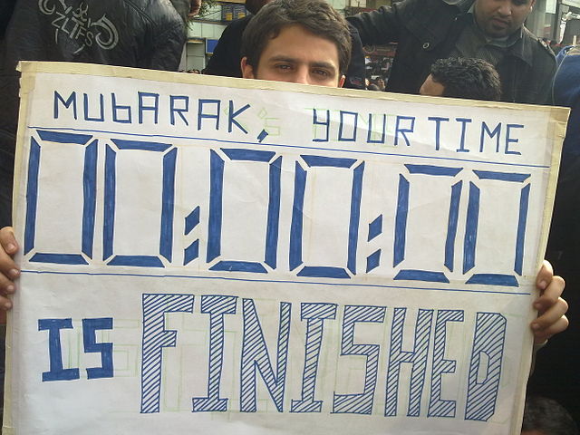 A young man carrying a card during the 2011 Egyptian Revolution in Tahrir Square saying "Mubarak Your Time is Finished"