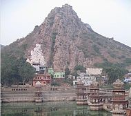 Daytime view of a more-or-less cone-shaped peak, perhaps reaching two hundred feet above the surrounding terrain. It is sparsely covered in poor-looking shrubs and trees; it otherwise reveals only an aspect of heated naked rock. Assorted parti-coloured blocky concrete buildings reach down from its lower slopes to a temple tank in the near foreground, around which is arrayed the more rounded and ornate temple structures.