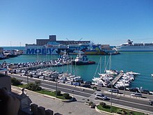 Udsigt over en havn med en bilbane i forgrunden, lystbåde i baggrunden og i baggrunden havet og en voluminøs færge