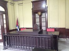 In this Havana courtroom, the professional judge sits in the chair at the center of the bench with the two lay judges at either side. Courtroom at Tribunal Municipal Popular 10 de Octubre, Havana, Cuba.jpg