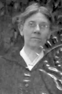 A middle-aged white woman wearing glasses, a light blouse, and a dark jacket