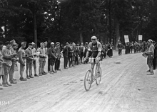 Черно-белая фотография, показывающая велосипедиста в гонке со зрителями на обочине дороги.