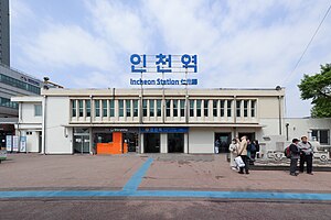 The front of the Incheon Station. The building is two stories tall and has a large blue sign on top reading Incheon Station in Korean, English, and Japanese. The pavement in front of the station has blue tactile paving marking the route into the station. There are two entrance doors to the station, and a small statue of a steam train can be seen to the right. It is a bright day and the sky is slightly cloudy.