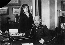 Une femme voilée en tenue de deuil se tient debout derrière un homme chauve et moustachu en tenue militaire assis à un bureau.