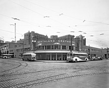 Black and white photo of corner building on a semi deserted street with round facade, sign saying Northland Greyhound, and two vertical signs reading Greyhound, surrounded by buses, Pantages Vaudeville and other buildings visible in rear