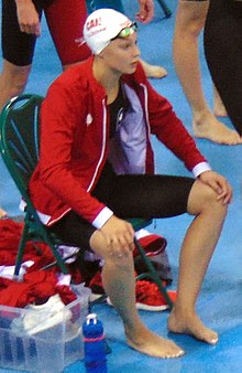 Penny Oleksiak in a swimsuit and jacket, seated on a bench by the Olympic pool.