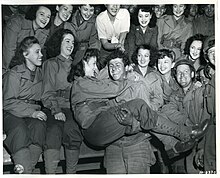 Female USO troupe entertaining an American soldier, 1944. SC 191837-S - A GI gets an armful of beauty as a U.S.O. camp show puts on their first entertainment for American troops on the Continent (50064074473).jpg