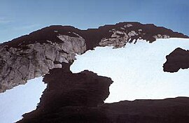 Изрезанная гора, покрытая темными скалами на вершине и склонах. На переднем плане вырисовываются темные скалы и участки ледникового льда.