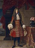 Oil painting of a white man with a large, curled wig of a medium brown. He is wearing a red coat ostentatiously decorated with cording of horizontal, diamond-shaped designs. The coat is fitted to the hips where it flares out and stops at the knees. The sleeves are long and fitted with lace around the cuffs. Lace also falls over the neck of the coat. Underneath the coat, the man is wearing black stockings and black shoes. He is holding a walking stick and standing next to a table covered in red and gold cloth.