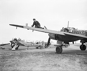 A photograph of a single–engined and a larger, twin-engined aircraft on the ground. Two men are working on the wing of the larger aircraft. The engine–covers have been removed from the smaller aircraft and four men are working on its engine