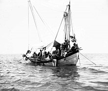 Cliché noir et blanc. Un scaphandrier sur le flanc d'un bateau. Un autre, sans casque de plongée, à cheval sur le bastingage. Sept hommes d'équipage d'un bateau à voile ancré.